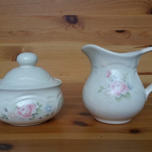 Peut inclure: Sucrier en céramique crème avec couvercle et pichet assorti. Les deux pièces présentent un motif floral avec des roses roses et bleues. Le sucrier a un bord festonné.
