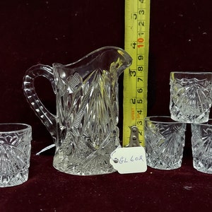 May include: A set of clear glass drinking glasses and a pitcher with a cut glass design. The pitcher has a handle and a wide mouth. The glasses are all the same size and shape. The set is on a red velvet surface.