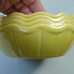 Puede incluir: Un cuenco de cerámica amarilla con borde ondulado. El cuenco tiene un acabado liso y brillante y una forma redondeada. El cuenco mide aproximadamente 15 cm de diámetro y probablemente se utiliza para servir alimentos o como pieza decorativa.