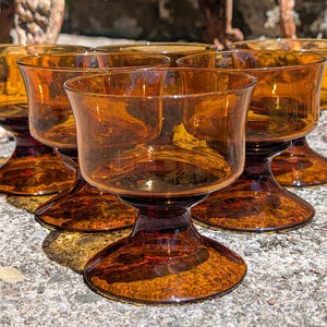 May include: A set of six amber-colored glass dessert bowls, each featuring a wide, shallow basin supported by a thick, sturdy pedestal base. The glassware is crafted from translucent, warm-toned glass with a smooth, polished finish throughout.
