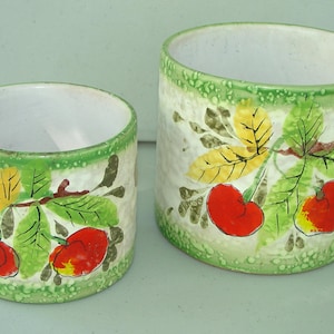 Puede incluir: Dos macetas de cerámica blanca con diseños florales verdes y amarillos y cerezas rojas. La maceta más grande mide 15 cm de alto y la maceta más pequeña mide 10 cm de alto.