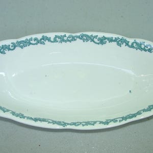 May include: Oval-shaped white serving dish with a teal floral design around the rim. The dish has a scalloped edge and is suitable for serving food. The teal design adds a decorative touch to the dish.