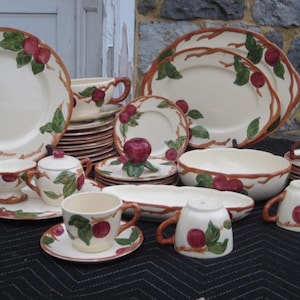 Pode incluir: Uma coleção de louças de cerâmica creme com um design de maçã vermelha e folhas verdes. O conjunto inclui pratos, tigelas, xícaras, pires e travessas. A borda marrom acentua as bordas das peças.