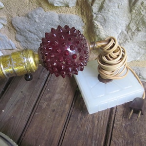 May include: A vintage lamp featuring a clear lightbulb, a brass-coloured socket, and a deep red, spiky glass shade. The lamp rests on a white square base with a coiled tan cord and a brown plug. The background is a wooden surface and a stone wall.