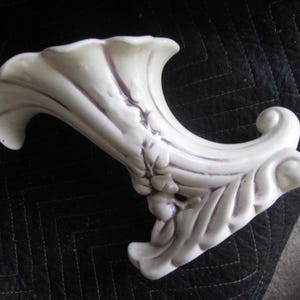 May include: A cream-colored ceramic cornucopia vase featuring a flared, ruffled opening and a curved, tapered body. The exterior surface is decorated with molded floral accents and subtle purple detailing along the recessed ridges and edges.