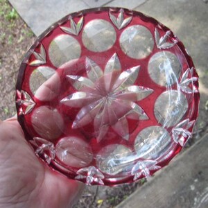 May include: A red glass dish with a central starburst design and circular patterns around the rim. The dish is held in a hand, highlighting its detailed cut glass. The item is likely a decorative piece, possibly for display.