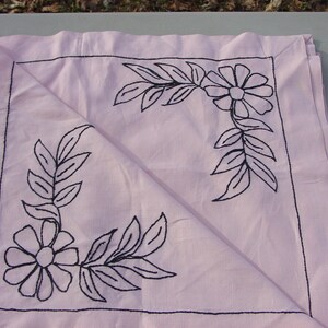 May include: A pink fabric square with black embroidered floral designs in the corners. The fabric is folded in half, showing the embroidery on both sides.