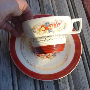 Puede incluir: Una taza de té blanca con un patrón floral rojo y dorado y un borde rojo. La taza de té está sentada sobre un platillo a juego con un borde rojo y un patrón floral dorado.