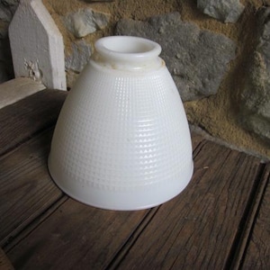 May include: A white, textured lampshade with a flared shape. The lampshade has a grid-like pattern and a slightly aged appearance. It sits on a wooden surface, suggesting a vintage or antique aesthetic.