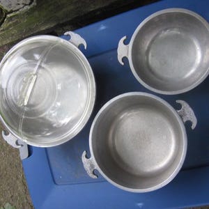 May include: A set of three round metal cooking pots with textured side handles. One pot includes a clear glass lid with a central handle. The pots have a matte silver finish and are arranged in a triangular formation on a flat surface.