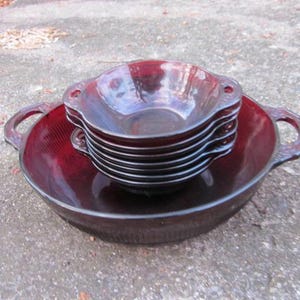 May include: A set of ruby red glass bowls. The set includes a large bowl with handles and a stack of smaller bowls. The bowls have a textured design and are arranged on a concrete surface.