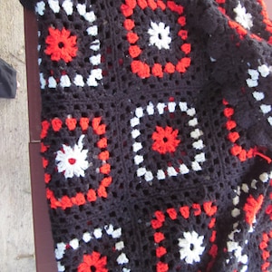 May include: Black crochet blanket with red and white squares. The blanket is made of a thick yarn and has a textured surface.