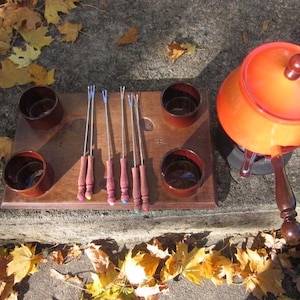 May include: An orange fondue pot with a wooden handle and a matching lid sits next to a wooden serving tray. The tray holds six fondue forks and four small brown bowls. The scene is set outdoors with autumn leaves.