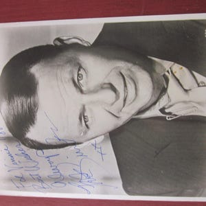 Peut inclure: Photographie en noir et blanc d'un homme en costume, souriant et regardant la caméra. La photographie est signée à l'encre bleue. L'homme a les cheveux foncés et porte une chemise à col.