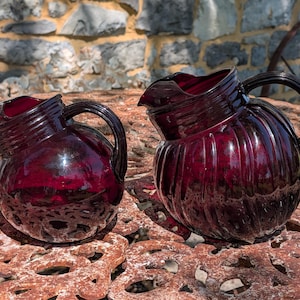 May include: Two antique, ruby-red glass pitchers with ribbed detailing and curved handles sit on a weathered, rusty metal surface. The pitchers have a bulbous shape and a spout for pouring. The background features a stone wall and a partial view of a metal wheel.