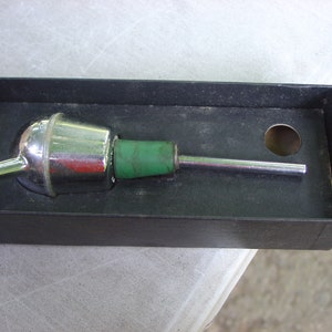 Puede incluir: Un vertedor de botella de licor plateado y verde en su caja original. El vertedor tiene una parte superior redonda y bulbosa y un pico largo y delgado.