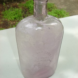 Puede incluir: Una botella de vidrio morado claro vintage con un cuerpo redondeado y un cuello largo. La botella tiene una apariencia descolorida y texto en relieve que dice "FRANK SABO 862 S MAIN ST PHILLIPSBURG NJ". La botella es probablemente una antigüedad.