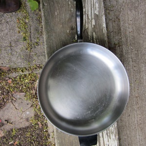 May include: A silver frying pan with a black handle. The pan is sitting on a wooden surface with white paint.