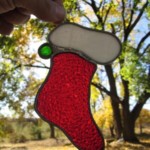 Peut inclure: Une décoration de Noël en verre teinté représentant une chaussette rouge et blanche avec une perle verte au sommet.