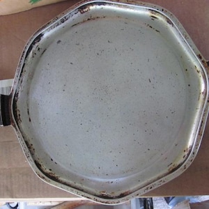 May include: A silver-toned, hexagonal metal cooking pan with a light-colored interior surface featuring dark speckling. The pan includes two black, stylized handles on opposite sides, showing signs of wear and discoloration along the edges.