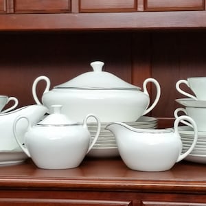 Peut inclure: Un ensemble de vaisselle en porcelaine blanche avec une bordure argentée. L'ensemble comprend un bol de service, un pot à crème, un sucrier, une saucière et un ensemble d'assiettes et de bols.