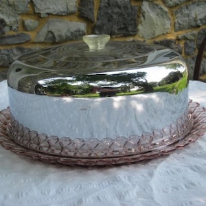 Op de afbeelding: Een vintage taartstandaard met een spiegelende chromen koepel en een roze glazen basis. De deksel reflecteert de omgeving. De basis heeft een decoratieve, geschulpte rand. De transparante knop bovenop is lichtgeel.