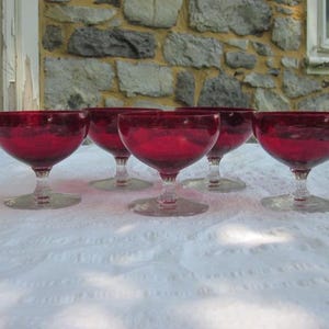 May include: Six vintage ruby red glass goblets. Each goblet features a rounded bowl, a clear stem, and a circular base. The glasses are arranged on a white textured surface, with a stone wall in the background.