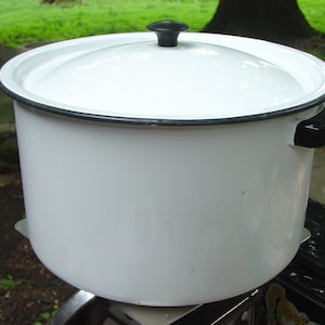 May include: A white enamel cooking pot with a lid. The pot has a black rim and handles. The lid has a black knob. The pot is sitting on a metal surface, with a green background.