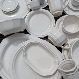 May include: An assortment of white ceramic dishware, including plates, saucers, bowls, and cups. The pieces have a classic design with subtle embossed detailing. The arrangement suggests a complete dinnerware set, perfect for everyday use or special occasions.