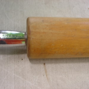 May include: A wooden rolling pin with a light brown finish and a silver metal handle. The rolling pin is designed for baking and features a cylindrical shape for even dough rolling. The handle is attached to the wooden barrel.