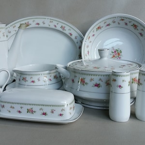 May include: A white porcelain dinnerware set with a floral pattern. The set includes a teapot, serving platters, bowls, a butter dish, salt and pepper shakers, and a covered casserole dish. The floral design features pink and yellow roses with green leaves.