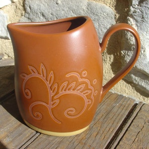 Puede incluir: Una jarra de cerámica marrón con una base de color crema y un diseño floral. La jarra tiene un asa y una boca ancha.