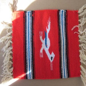 Op de afbeelding: Een felrood geweven textiel met een centraal wit en blauw vogelontwerp. Het textiel heeft verticale strepen in zwart, wit en blauw. Crèmekleurige franjes sieren de randen en geven dit decoratieve stuk textuur.