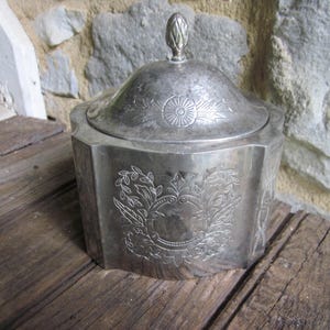 May include: A silver-colored, lidded container with an octagonal base. The lid is topped with a decorative finial. The container features engraved floral and circular designs. The container is sitting on a wooden surface.