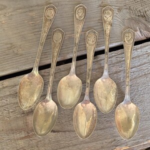 May include: Six antique silver spoons with detailed handles featuring a portrait design. The spoons are arranged on a weathered wooden surface, showcasing their aged patina and intricate craftsmanship. A vintage silverware set.