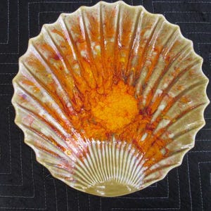 May include: A ceramic, shell-shaped serving dish with a scalloped edge. The dish features a vibrant orange and yellow center that fades into a cream-colored exterior. The design has a radial pattern, with vertical lines extending from the center to the edge.