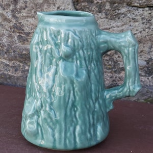 May include: A green ceramic jug with a tree bark texture. The jug has a handle that is shaped like a tree branch.