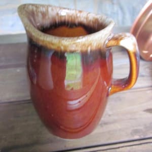 Peut inclure: Une cruche en céramique marron avec une anse. La cruche a une finition brillante avec un bord marron plus foncé et une bande marron plus claire. L'intérieur est marron plus foncé. La cruche est sur une surface en bois.