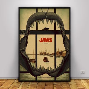 Peut inclure: Une affiche de film encadrée pour Jaws représentant la silhouette d'une grande mâchoire de requin sombre encadrant une scène de port avec des bateaux et des bâtiments. Le titre JAWS est imprimé en lettres rouges grasses au centre sur fond beige.