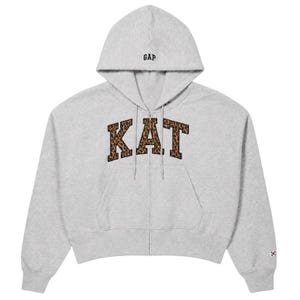 May include: A light grey zip-up hoodie featuring the word KAT in a brown and black leopard print pattern across the chest. The garment includes a hood with drawstrings, a front zip closure, and the GAP logo embroidered in black on the hood.