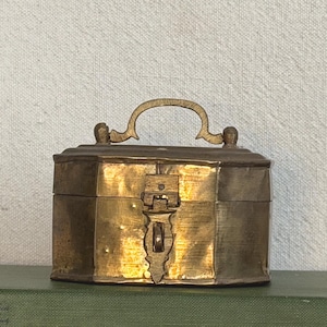 May include: A small, hexagonal brass box with a hinged lid and a curved handle on top. The box features a central latch mechanism on the front panel. The metal surface has a polished, reflective finish with visible variations in tone and texture.