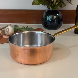 May include: A round saucepan with a polished copper exterior and a stainless steel interior. The cookware features a long, slender, gold-toned metal handle extending from the side. The pan has a smooth, cylindrical body and a flat base.