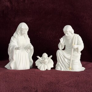 May include: A three-piece white ceramic nativity set featuring a kneeling figure with a head covering, a central infant figure resting in a floral-shaped cradle, and a kneeling bearded figure holding a staff with one hand raised in a gesture.