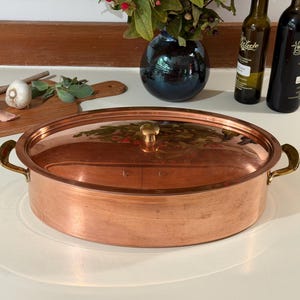 May include: An oval copper roasting pan with a matching lid and two brass side handles. The pan features a polished metallic finish and a central brass knob on the lid. The design is simple and functional, showcasing the warm, reddish-brown tones of the copper material and the contrasting gold-colored brass accents on the handles and lid.