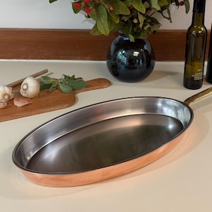 May include: An oval-shaped copper frying pan with a polished stainless steel interior and a long, slender brass handle. The exterior features a warm, metallic copper finish, while the interior surface is smooth, reflective, and silver-toned.