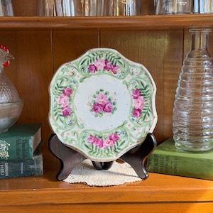 May include: A decorative ceramic plate with a scalloped edge, featuring a central cluster of pink roses surrounded by green fern-like foliage and additional rose motifs along the border, displayed on a dark brown wooden stand.