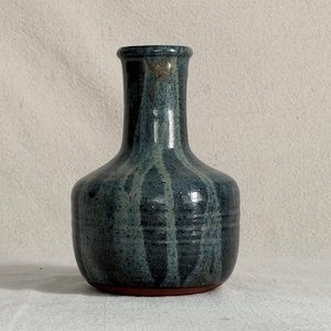 May include: A ceramic vase with a bulbous base and a narrow neck, featuring a glossy glaze in shades of deep blue and grey with vertical drip patterns. The bottom edge reveals a raw, unglazed terracotta-colored clay rim at the base.