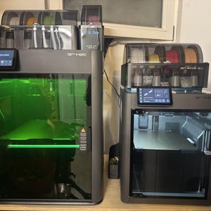 Op de afbeelding: Twee Bambu Lab 3D-printers, een X1C en een X1 Carbon, staan naast elkaar. Beide apparaten zijn voorzien van gesloten behuizingen met glazen deuren, AMS-filamenttoevoersystemen aan de bovenzijde en geïntegreerde touchscreens voor de status.