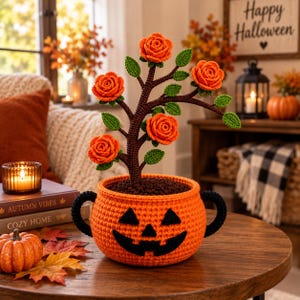 Puede incluir: Una maceta de ganchillo naranja con forma de calabaza, cara de jack-o'-lantern negra y dos asas negras. Un árbol de ganchillo marrón crece de la maceta, con cuatro rosas naranjas y varias hojas verdes de ganchillo en sus ramas.