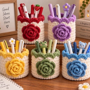 Puede incluir: Cinco portalápices cilíndricos de ganchillo en color crema con un borde superior festoneado en rojo, morado, amarillo, verde y azul. Cada soporte presenta una flor de ganchillo a juego en la parte frontal y contiene varios bolígrafos.